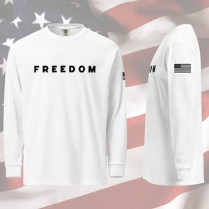以下が含まれることがあります： 胸に黒字で「FREEDOM」とプリントされた白の長袖シャツ。左袖には小さなアメリカ国旗のグラフィックがあります。背景にはアメリカ国旗が描かれています。