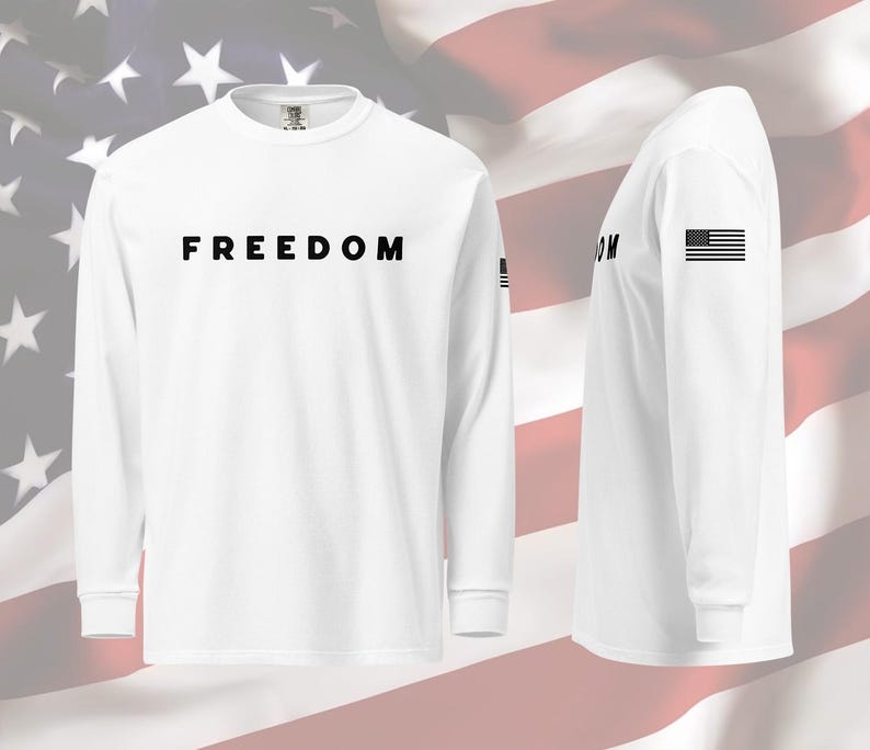 以下が含まれることがあります： 胸に黒字で「FREEDOM」とプリントされた白の長袖シャツ。左袖には小さなアメリカ国旗のグラフィックがあります。背景にはアメリカ国旗が描かれています。