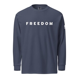 以下が含まれることがあります： 胸に白で「FREEDOM」の文字がプリントされた、ネイビーブルーの長袖シャツ。右袖には小さな白と青のグラフィックがあります。シャツはクルーネックで、ゆったりとしたフィット感です。