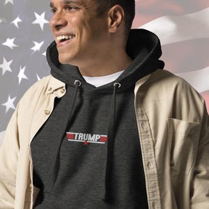 May include: A dark gray hooded sweatshirt with a red, white, and blue graphic of the word "TRUMP" with a star underneath. The sweatshirt is being worn by a person in front of an American flag.