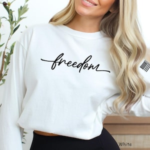 Könnte beinhalten: Ein weißes Langarm-Sweatshirt mit dem Wort "freedom" in schwarzer Schrift auf der Brust. Die amerikanische Flagge ist auf dem rechten Ärmel aufgedruckt. Das Wort "White" ist unten rechts im Bild zu sehen.