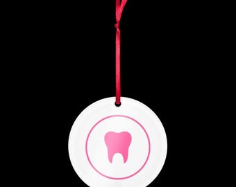 May include: A white, round ornament with a pink tooth graphic inside a pink circle. A red ribbon is attached for hanging. The ornament is likely for dental professionals or as a dental-themed gift.