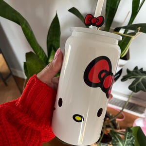May include: White reusable cup with a Hello Kitty design. The cup has a red bow and a straw.