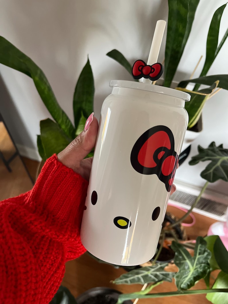 May include: White reusable cup with a Hello Kitty design. The cup has a red bow and a straw.