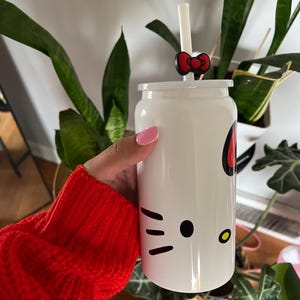 May include: White Hello Kitty themed reusable water bottle with a black Hello Kitty face, a red bow, and a clear straw. The bottle is being held by a hand wearing a red sweater.