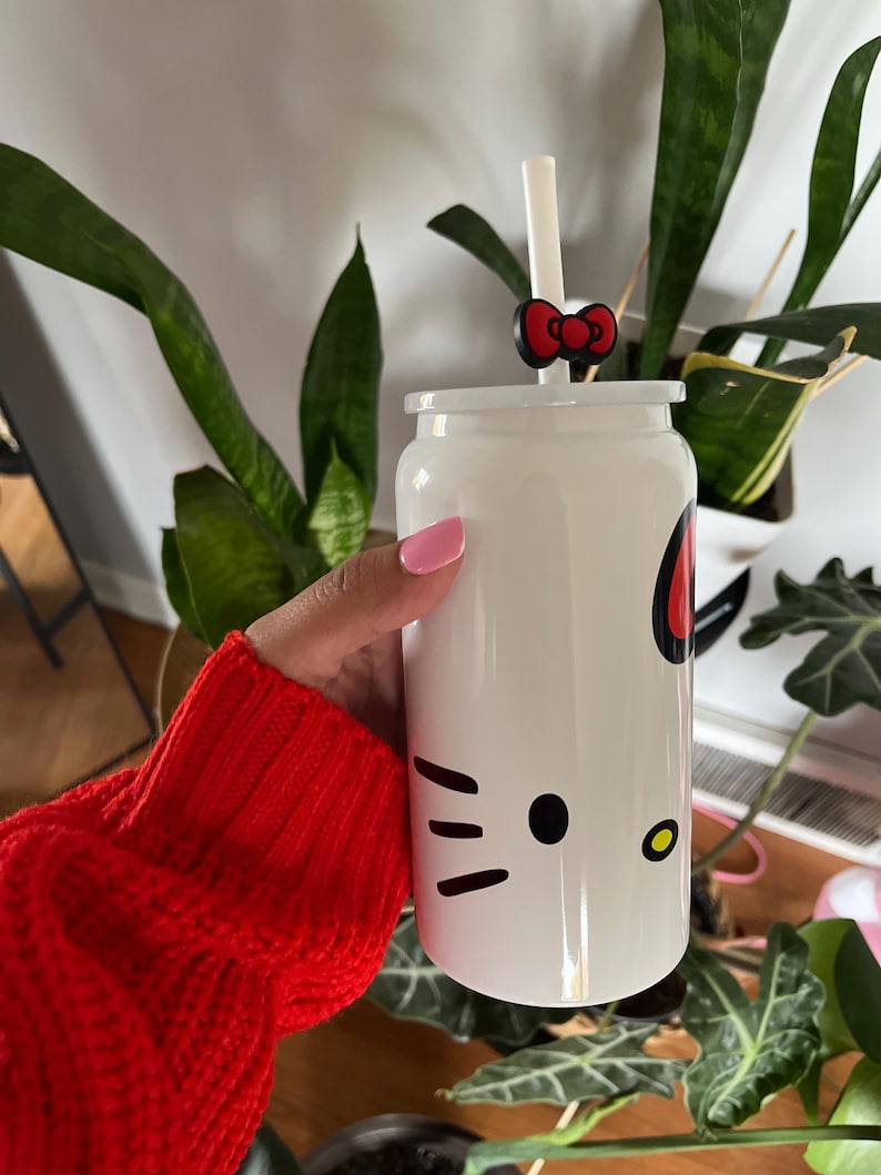 May include: White Hello Kitty themed reusable water bottle with a black Hello Kitty face, a red bow, and a clear straw. The bottle is being held by a hand wearing a red sweater.