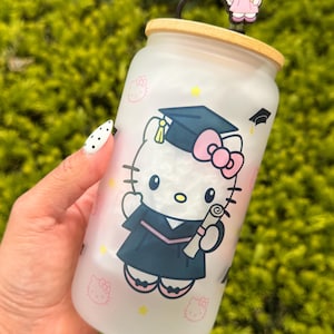 May include: A frosted glass tumbler featuring a graduation-themed Hello Kitty design. The tumbler has a wooden lid and a straw with a Hello Kitty charm. The design includes Hello Kitty in a graduation gown, mortarboard, and diploma, with pink and yellow accents.