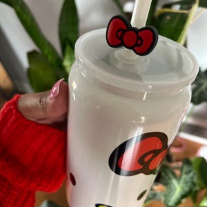 May include: White ceramic tumbler with a red and black Hello Kitty design. The tumbler has a straw and a red bow on top.