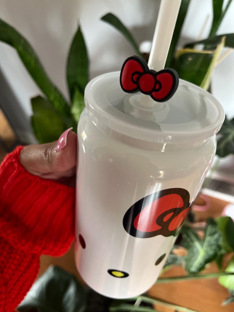May include: White ceramic tumbler with a red and black Hello Kitty design. The tumbler has a straw and a red bow on top.