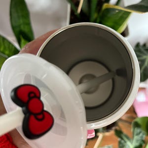 May include: White stainless steel tumbler with a red Hello Kitty bow on the lid. The tumbler has a straw and a clear lid.