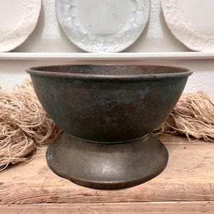 May include: A round, deep metal bowl with a weathered, dark patina featuring shades of brown, green, and copper. The bowl is supported by a wide, circular pedestal base that matches the aged, oxidised finish of the main vessel body.