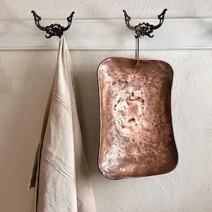 Peut inclure: Un plateau rectangulaire de couleur cuivre avec une poignée est suspendu à un crochet décoratif en métal foncé. Une serviette beige est drapée sur un crochet similaire à gauche. Le plateau a une finition vieillie, et les crochets sont montés sur une planche de bois peinte en blanc.
