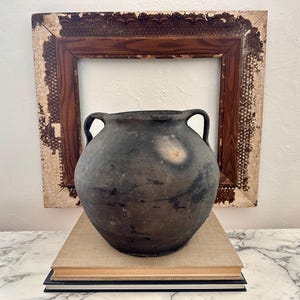 Peut inclure: Un pot en céramique gris foncé de style antique avec deux anses, posé sur une pile de livres. Le pot a un aspect vieilli avec une tache de couleur claire. Derrière, un cadre en bois vieilli.