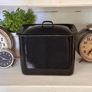 Peut inclure: Une boîte à pain rectangulaire noire en émail avec un couvercle et une poignée. La boîte à pain est sur une surface blanche, avec deux réveils vintage et une petite plante en pot en arrière-plan. Les horloges ont des cadrans blancs et noirs.