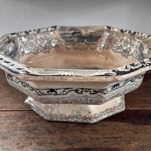 May include: An antique, octagonal ceramic bowl with a pedestal base. The bowl has a cream-coloured interior and is decorated with black floral and scrollwork patterns along the rim and exterior. The bowl is displayed on a wooden surface.