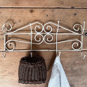 Peut inclure: Un porte-manteau mural en métal blanc avec une finition vieillie, doté de volutes décoratives et de plusieurs crochets. Un petit panier tressé marron foncé est suspendu à un crochet, et un tissu texturé clair est suspendu à un autre.