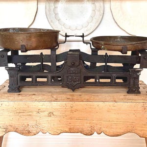 May include: Vintage black and bronze balance scale with two copper pans. The scale is marked "SARAY 5 KG" and is placed on a rustic wooden table. Decorative plates are in the background.