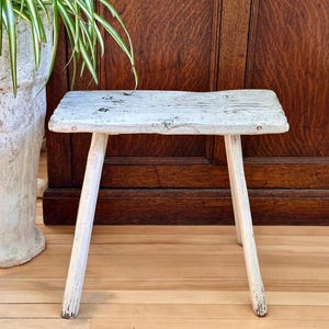 Peut inclure: Un petit tabouret rectangulaire en bois avec une finition peinte en blanc vieilli. Le tabouret présente une surface supérieure plate et texturée avec un grain de bois visible et quatre pieds robustes légèrement inclinés.