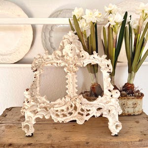 Può includere: Cornice decorativa ornata bianco sporco con finitura anticata, appoggiata su una superficie in legno rustico. La cornice ha un design dettagliato a volute. Fiori bianchi in un piccolo vaso sullo sfondo.