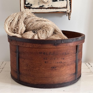 Op de afbeelding: Een ronde houten schepelmand met donkere metalen banden en klinknagels, gevuld met een grote bundel lichtbruin natuurvezelgaren. De zijkant van de mand is bedrukt met de tekst HALF BUSHEL, THOS. PEEDS, ST. THOMAS en een klein logo.
