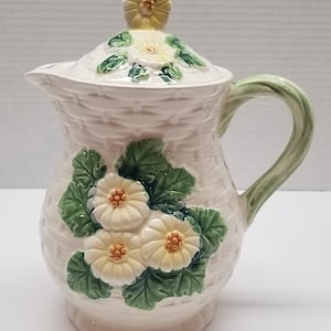 May include: A white ceramic pitcher with a lid, featuring a woven basket pattern and floral accents. The pitcher has a green handle and is decorated with white and yellow flowers.