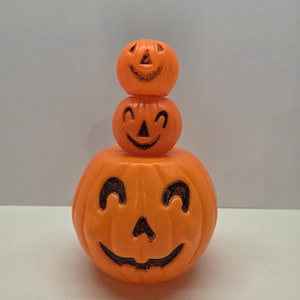 May include: Three orange plastic jack-o'-lanterns stacked on top of each other. Each pumpkin has a carved smiling face with black accents. The pumpkins are a classic Halloween decoration.