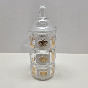 May include: A clear, three-tiered glass jar with a lid and handle. Each tier features a gold floral design. The jar is likely used for storing small items or as a decorative piece. The design is vintage and elegant.