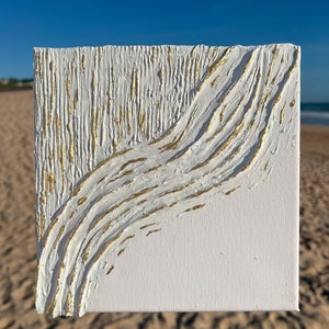 以下が含まれることがあります： テクスチャ加工された白い塗料と金色のアクセントが施された正方形の白いキャンバスアート。このアートワークは、左側にテクスチャ加工された垂直パターン、右側に流れるような斜めの金色のデザインが特徴です。キャンバスはビーチと青い空を背景に保持されています。