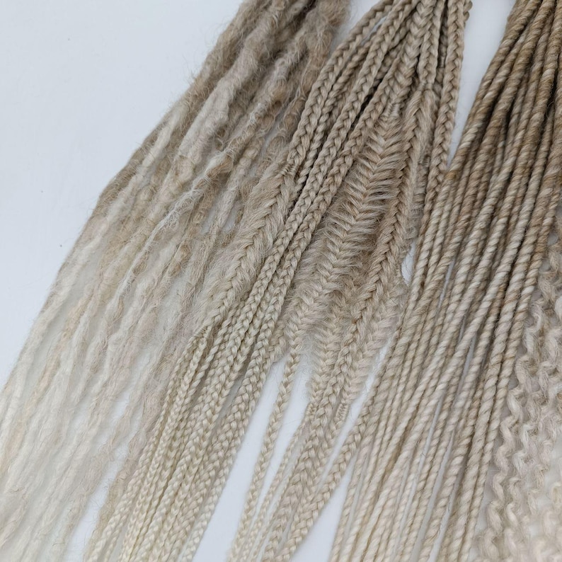 May include: A collection of blonde synthetic dreadlocks in various styles, including straight, braided, and twisted. The dreadlocks are ombre, transitioning from a light blonde to a darker blonde.