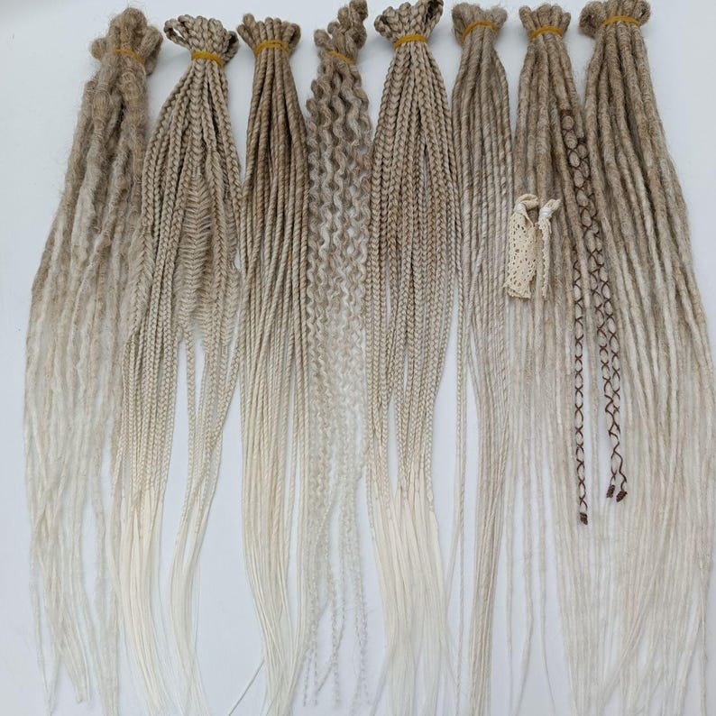 May include: A collection of 10 synthetic dreadlocks in various styles, including straight, wavy, and curly. The dreadlocks are ombre, transitioning from a light brown to a white colour.