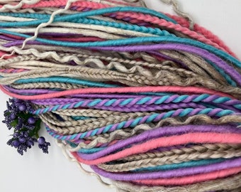 May include: A collection of colorful dreadlocks in shades of pink, purple, teal, and beige. The dreadlocks are arranged in a bundle, with some braided and others smooth. A small sprig of purple flowers is placed near the base of the bundle.