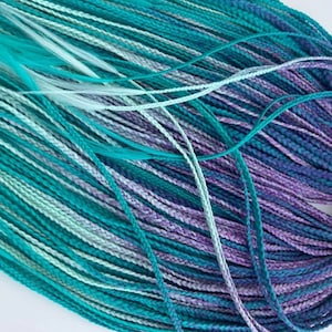 May include: A bundle of synthetic hair extensions in a variety of colours. The braids are a mix of teal, turquoise, light blue, and purple. The hair is tightly braided and tied together with a brown string.