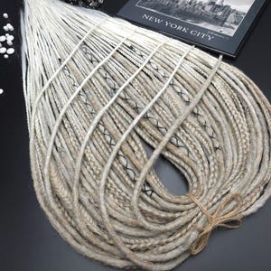 May include: A collection of blonde and light brown dreadlocks, secured with twine. The dreadlocks feature black thread detailing. A black and white photograph of New York City is in the background, with scattered white beads.