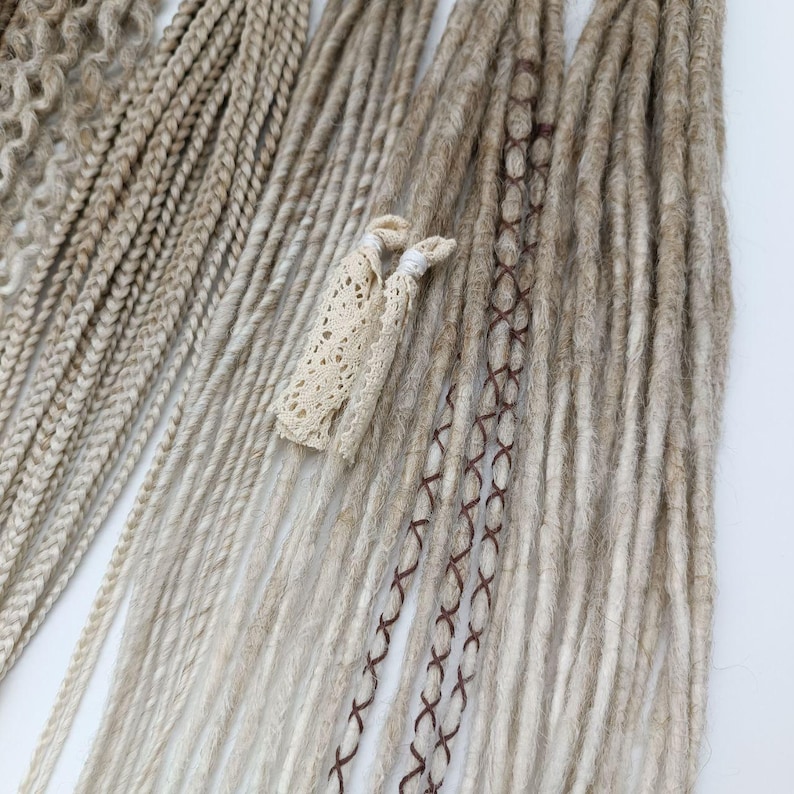 May include: A set of long, light brown synthetic dreadlocks with brown stitching. The dreadlocks are arranged in a fan shape and are tied together with a white lace ribbon.