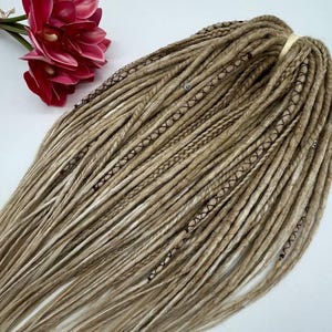 Puede incluir: Un conjunto de rastas sintéticas largas y rubias con costuras marrones. Las rastas están dispuestas en un paquete suelto.