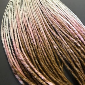 May include: A bundle of synthetic dreadlocks in shades of brown, pink, and white. The dreadlocks are ombre, with the lightest shade at the top and the darkest shade at the bottom.