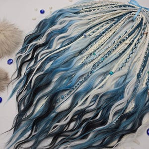 Könnte beinhalten: Eine Sammlung gewellter, synthetischer Dreadlocks in Blau-, Weiß- und Schwarztönen. Die Dreads sind mit Perlen und kleinen Metallringen verziert. Zwei flauschige, hellbraune Pompons und blaue Ziersteine sind ebenfalls vorhanden.