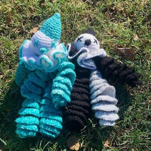May include: Two crocheted stuffed toys, one in turquoise and white, the other in black and white. The turquoise toy is shaped like a clown with a ruffled collar. The black and white toy is shaped like a panda with a ruffled collar.