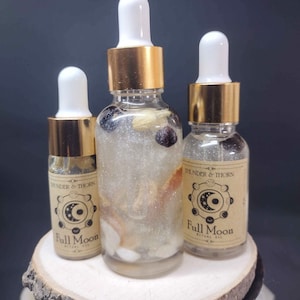 May include: Three glass bottles with gold lids and white droppers filled with a clear liquid and various crystals. The bottles have labels that say "Thunder & Thorn" and "Full Moon Ritual Oil".