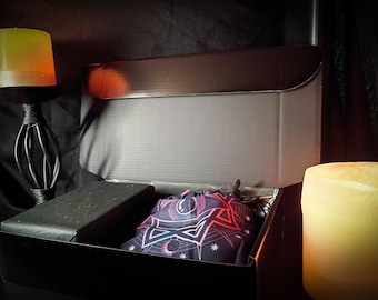 May include: A black box with an open lid, containing a black wrapped item and a drawstring bag with a celestial design in red, white, and blue. Two lit candles are on either side of the box, one yellow and one white.