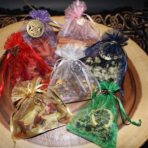 May include: Six small, sheer fabric bags filled with dried herbs and flowers. Each bag has a different colour and features a metal charm with a different symbol. The bags are arranged on a wooden surface.
