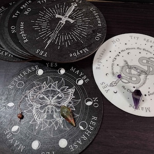 Könnte beinhalten: Ein Satz von sechs schwarzen Holz-Divinationsbrettern mit weißem Design. Jedes Brett hat ein anderes Design, darunter ein Schwert, ein Schmetterling und eine Schlange. Die Bretter werden mit einem Pendel verwendet, um Fragen zu beantworten.