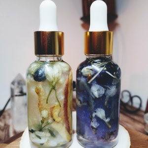 May include: Two glass bottles with gold lids and white droppers filled with a clear liquid and dried flowers. The bottle on the left contains white flowers and orange peels. The bottle on the right contains blue flowers.