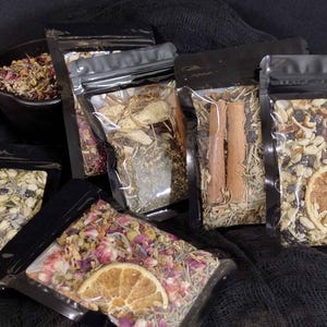 Può includere: Un assortimento di erbe e spezie essiccate in sacchetti trasparenti sigillati con cerniere nere. Alcuni sacchetti contengono fette d'arancia e bastoncini di cannella. È presente anche una ciotola nera piena di ingredienti simili.