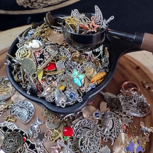 Könnte beinhalten: Eine schwarze Metallschale gefüllt mit einer Vielzahl von silbernen und goldenen Charms, darunter Sterne, Monde, Schlüssel und Herzen. Die Schale steht auf einer Holzoberfläche.