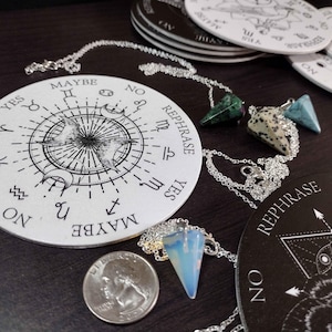 May include: A white circular pendulum board with the words "Rephrase", "Yes", "Maybe", and "No" around the edge. The board is decorated with astrological symbols. A silver chain with three different colored crystal pendants is hanging over the board.