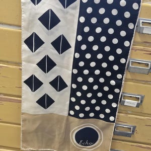 May include: A silk scarf with two distinct patterned sections. One side features black diamonds on a cream background, the other has white polka dots on navy. The scarf has a tan border and the word "Echo" is printed on it.