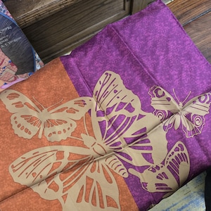 May include: A folded silk scarf featuring a butterfly design. The scarf has a color block design with orange and purple sections. The butterflies are tan and the scarf is made of a soft, silky material.