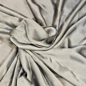 May include: A close-up of a draped, silvery-gray fabric with subtle patterns. The fabric has a silky sheen and is folded in soft, flowing curves. The background shows a glimpse of a red patterned fabric.