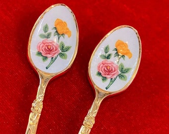 May include: Two gold-coloured spoons with floral designs. Each spoon features a white oval bowl with a pink and yellow rose illustration and green leaves. The handles have a decorative floral pattern.